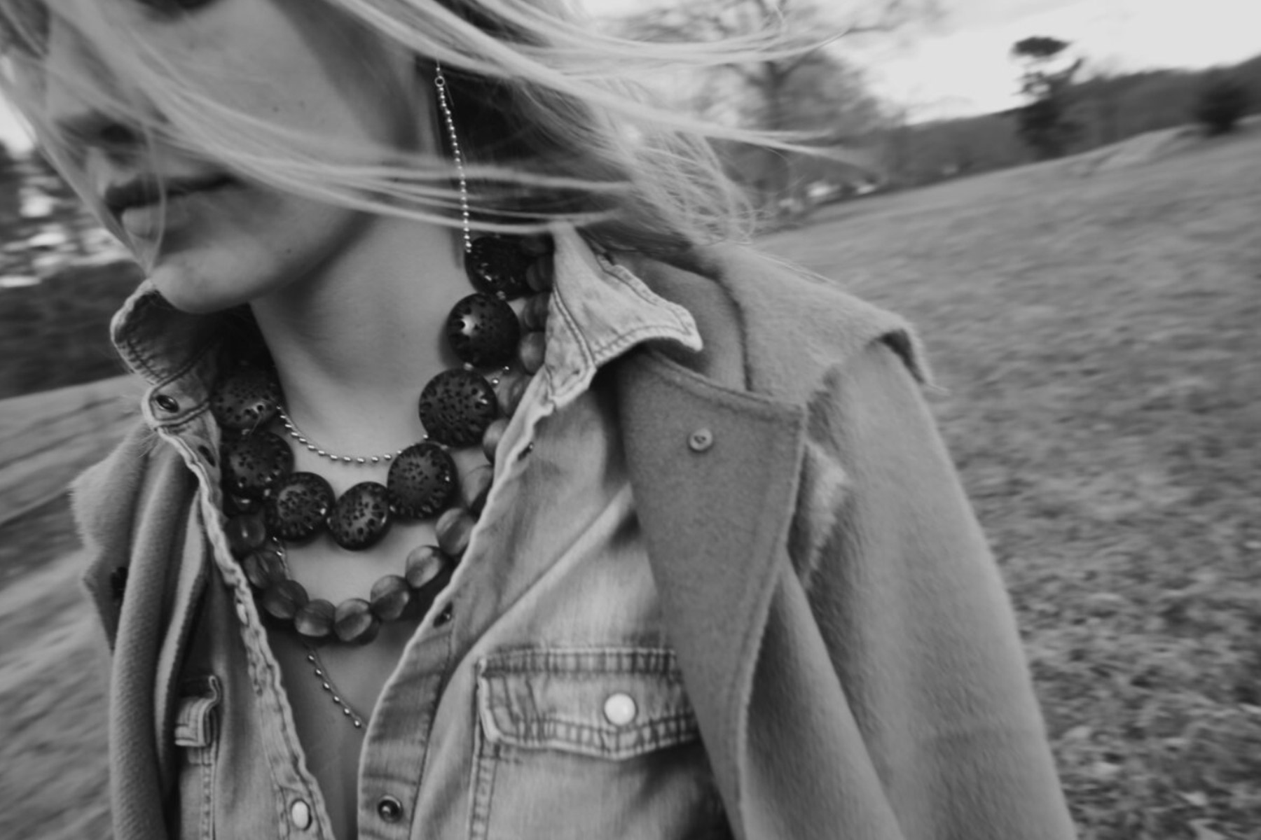 A black and white photo of a person wearing a denim jacket and layered necklaces, with long hair blowing in the wind. The background appears to be an open field with blurred trees and a cloudy sky. The image captures a sense of motion and casual style. Fearrington Village