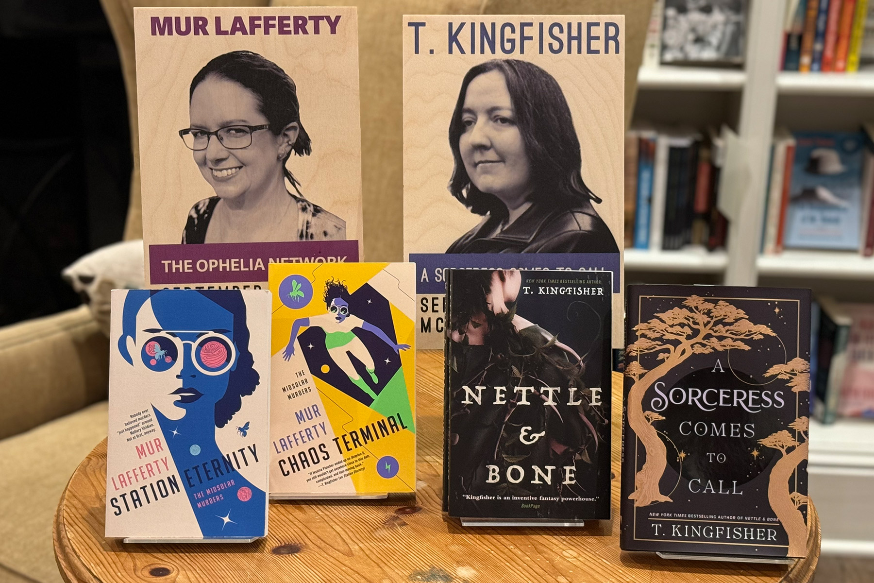 A display featuring books by Mur Lafferty and T. Kingfisher. Visible titles include "Six Wakes", "Staying Eternity", "Chaos Terminal" by Mur Lafferty, and "Nettle & Bone", "A Sorceress Comes to Call" by T. Kingfisher. The backdrop has images and names of the authors. Fearrington Village