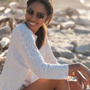 A woman with sunglasses smiles while sitting on a rocky beach. She is wearing a white and blue striped long-sleeve shirt. In the background, driftwood and rocks are illuminated by the sunlight. Fearrington Village