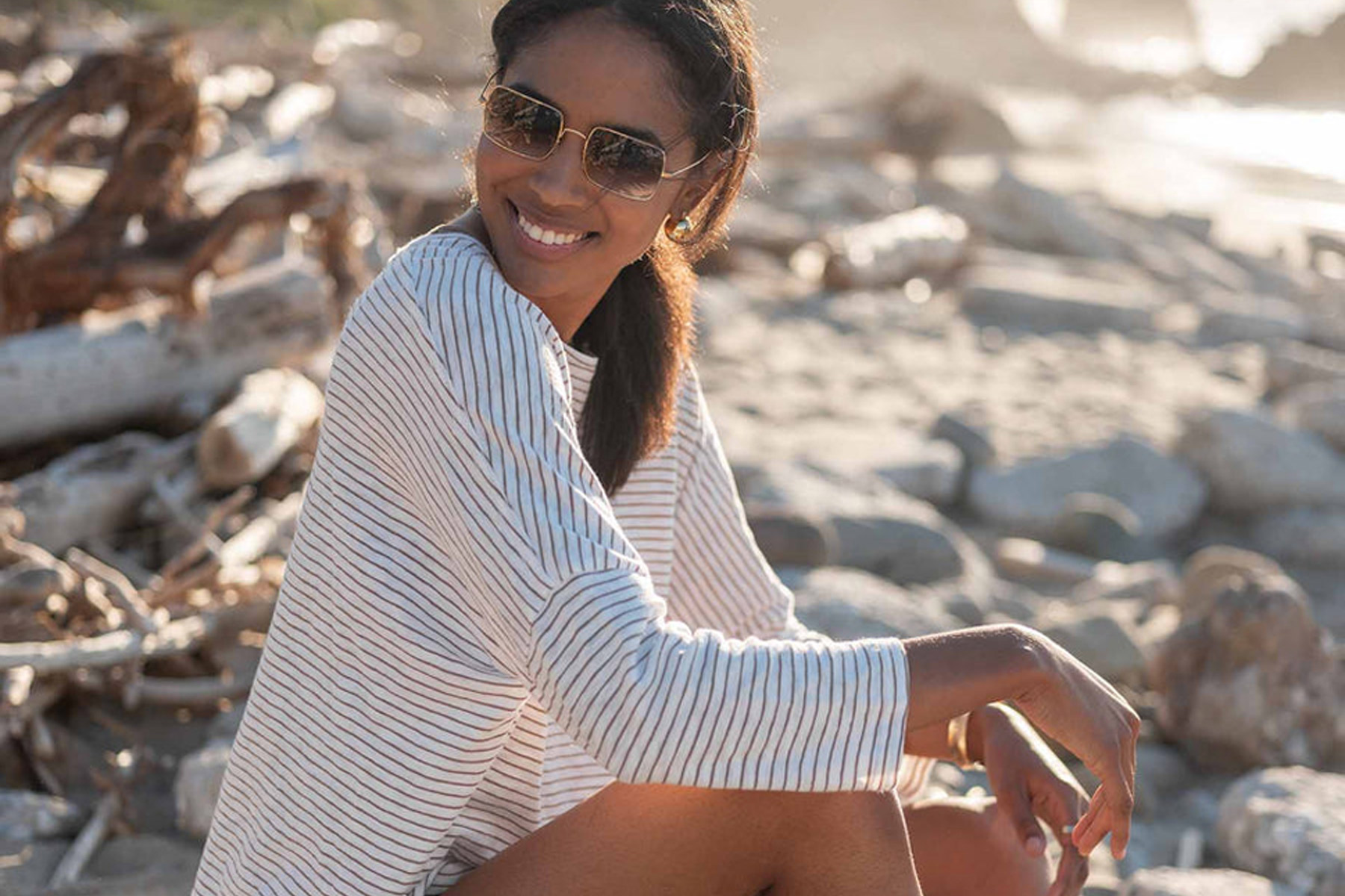 A woman with sunglasses smiles while sitting on a rocky beach. She is wearing a white and blue striped long-sleeve shirt. In the background, driftwood and rocks are illuminated by the sunlight. Fearrington Village