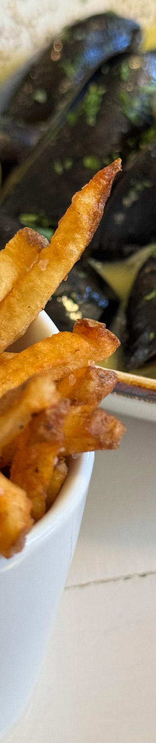 Close-up of a white cup filled with crispy, golden-brown fries in the foreground, with a plate of mussels garnished with green herbs slightly out of focus in the background. Fearrington Village