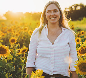 Natalie Bath. A woman with long blonde hair is smiling and standing in a field of sunflowers at sunset. She is wearing a light-colored, long-sleeved shirt, and the golden light of the setting sun illuminates the scene, giving it a warm, cheerful ambiance. Fearrington Village