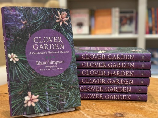 A display of the book "Clover Garden: A Carolinian's Piedmont Memoir" by Bland Simpson, featuring a purple cover with pine needles and pine cones. A stack of the books is situated on a wooden table with blurred shelves in the background. Fearrington Village