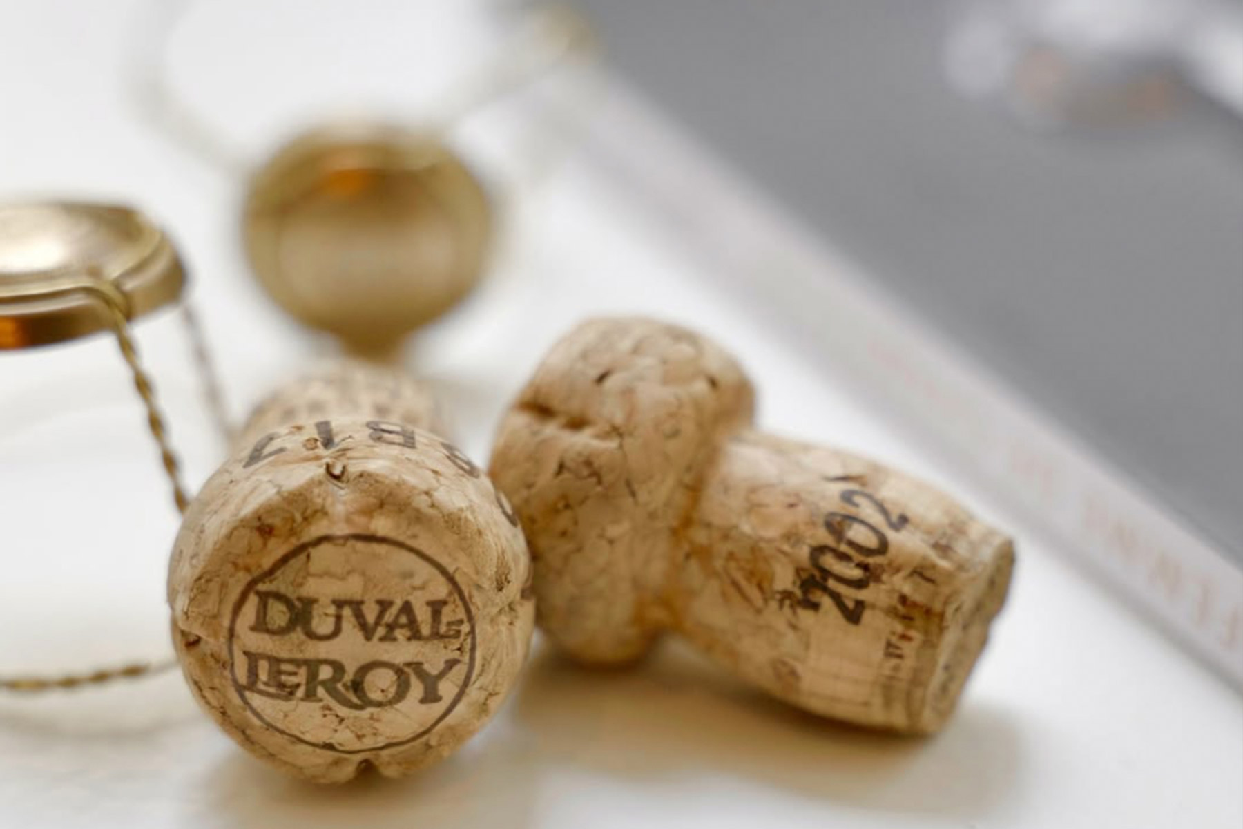 Two champagne corks, one branded "Duval-Leroy" and another marked "2002," are on a surface with a wire cage nearby. A blurred book or magazine is partially visible in the background. Fearrington Village