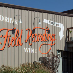 A building with the phrase "Drink The Way" in white and "Field Recordings Wine" in large orange cursive letters on the side. Outdoor seating is visible on a balcony. There are plants in the foreground. Fearrington Village