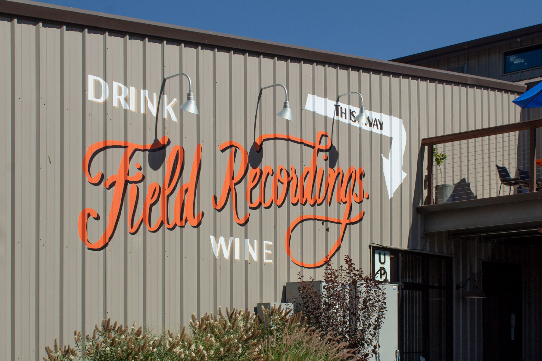 A building with the phrase "Drink The Way" in white and "Field Recordings Wine" in large orange cursive letters on the side. Outdoor seating is visible on a balcony. There are plants in the foreground. Fearrington Village