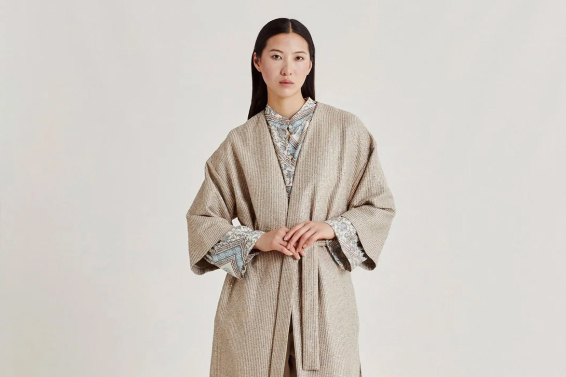 A person with long dark hair is wearing a beige, textured robe over a patterned shirt, posing against a plain background. The robe has wide sleeves and a belt tied at the waist. Fearrington Village