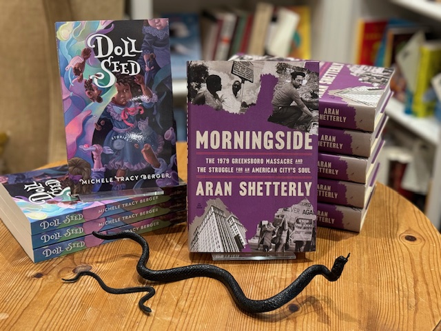 Books displayed on a wooden table: multiple copies of "Doll Seed" featuring a colorful cover with a woman's face and floral elements, and "Morningside" with a purple cover showcasing historical photographs. A black snake figurine decorates the table. Fearrington Village