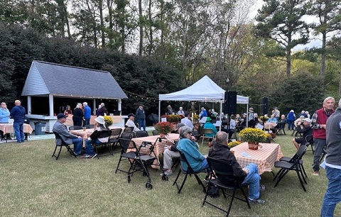 A group of people gathered outdoors for an event, sitting at tables with checkered cloths and flower centerpieces. There are two tents in the background, surrounded by trees and hedges, under a partly cloudy sky. Fearrington Village