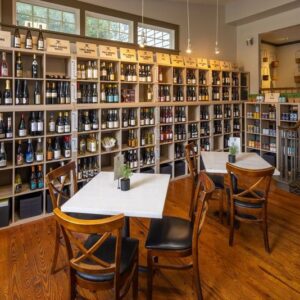 A cozy wine bar interior with wooden floors and neatly organized shelves filled with various wine bottles. Three small tables with chairs are arranged in the foreground, creating an inviting atmosphere for visitors. Fearrington Village