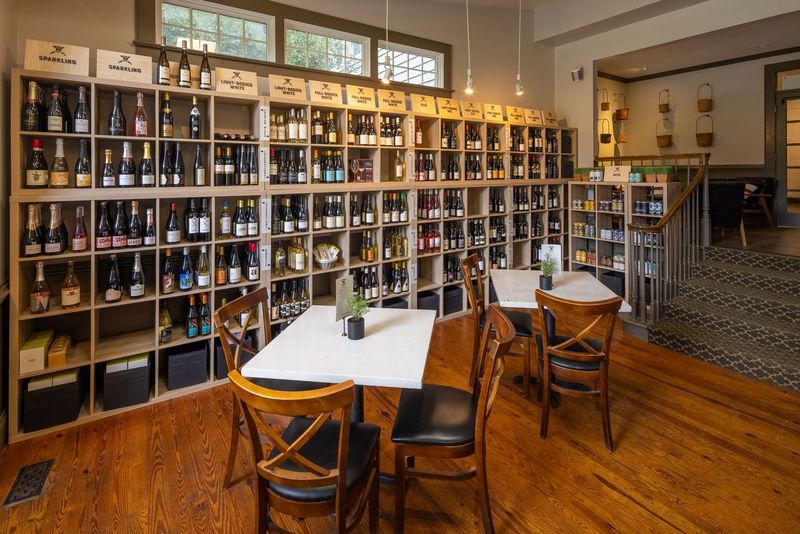 A cozy wine bar interior with wooden floors and neatly organized shelves filled with various wine bottles. Three small tables with chairs are arranged in the foreground, creating an inviting atmosphere for visitors. Fearrington Village