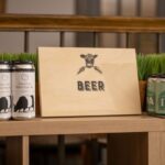 Cans of craft beer are displayed on a wooden shelf with a wooden sign in the middle featuring a cow illustration and the word "BEER." The left side has cans labeled "Farm Cow Kolsch-Style Ale," and the right side shows cans of IPA. Fearrington Village