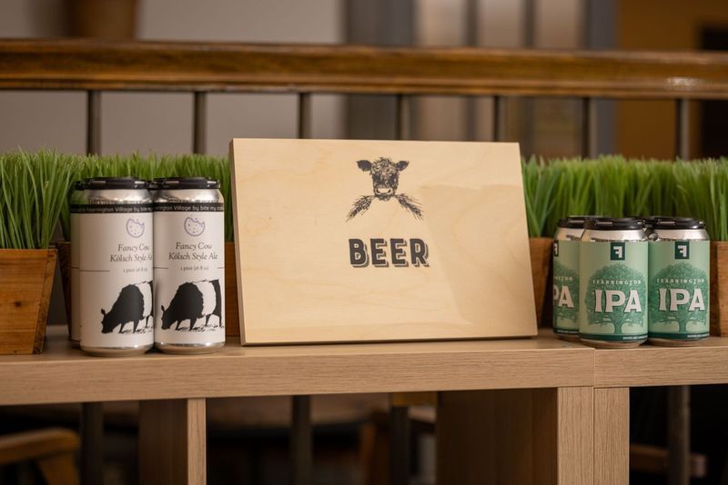 Cans of craft beer are displayed on a wooden shelf with a wooden sign in the middle featuring a cow illustration and the word "BEER." The left side has cans labeled "Farm Cow Kolsch-Style Ale," and the right side shows cans of IPA. Fearrington Village