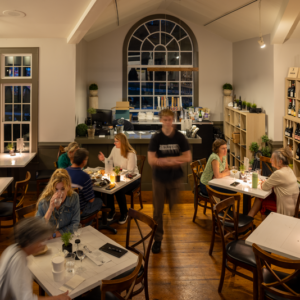 A cozy restaurant with wooden floors and shelves filled with wine bottles. Patrons sit at candlelit tables, enjoying their meals. A waiter is in motion near the center, while large windows add an elegant touch to the modern decor. Fearrington Village