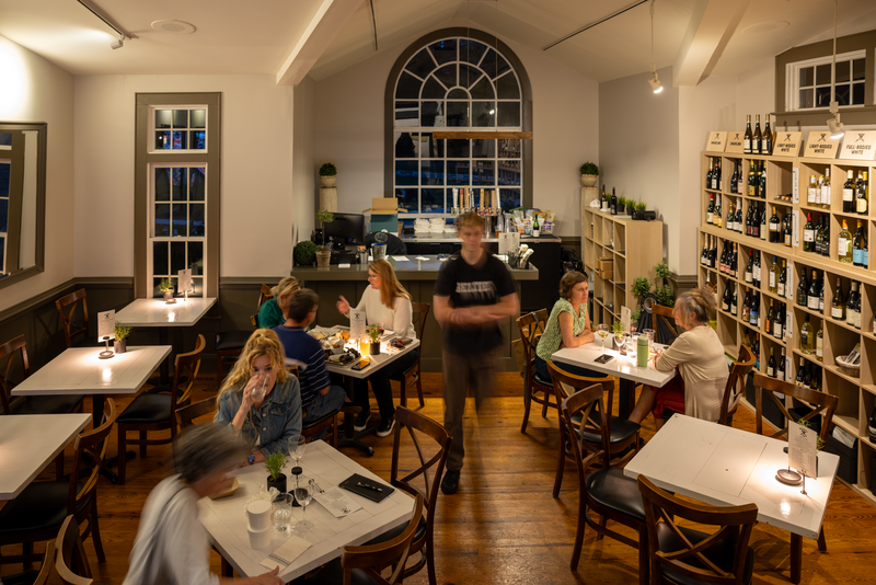 A cozy restaurant with wooden floors and shelves filled with wine bottles. Patrons sit at candlelit tables, enjoying their meals. A waiter is in motion near the center, while large windows add an elegant touch to the modern decor. Fearrington Village