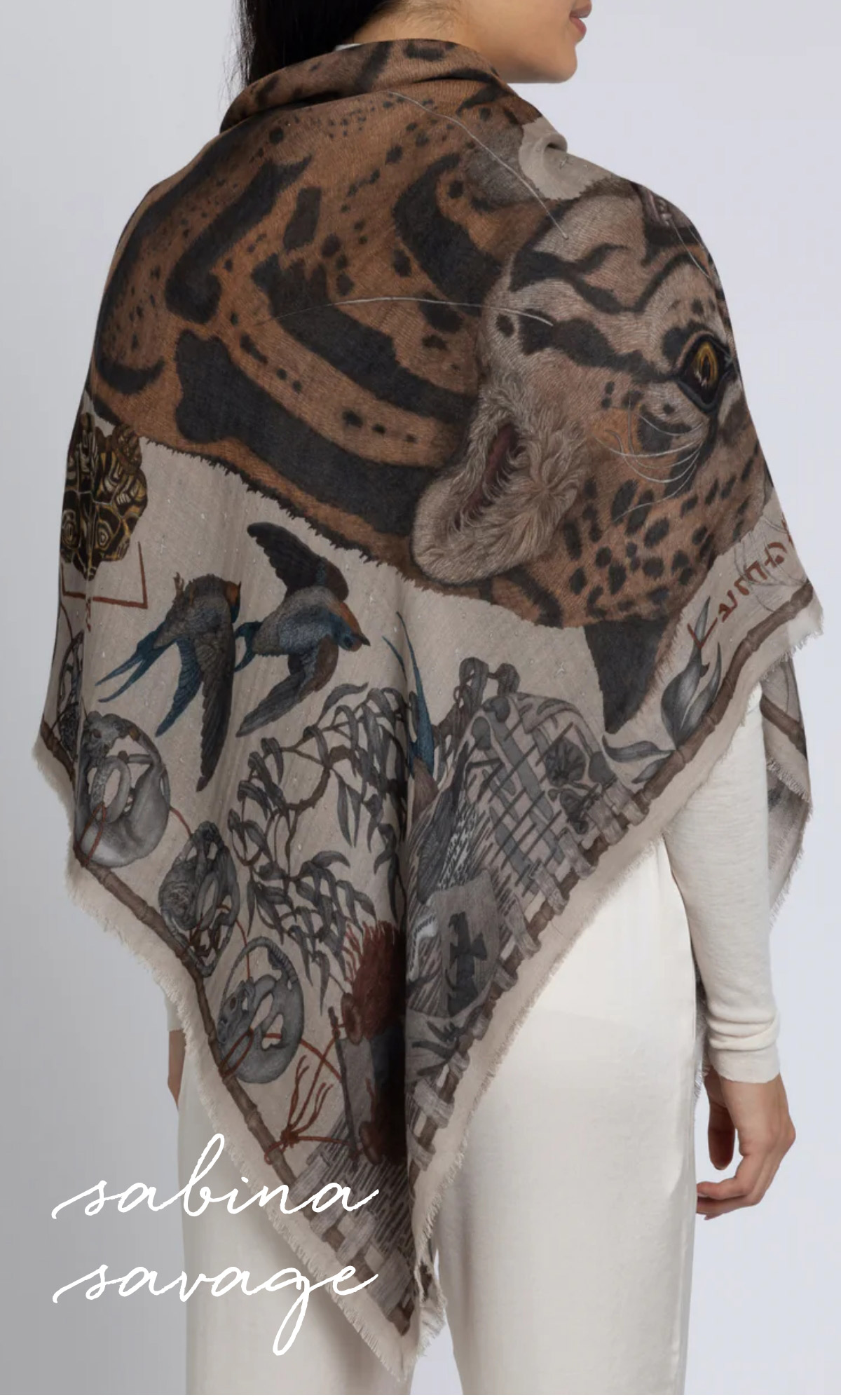 A person wearing a detailed scarf featuring images of a leopard, birds, and intricate designs. The scarf drapes over the shoulders with "Sabina Savage" written in the corner. Fearrington Village