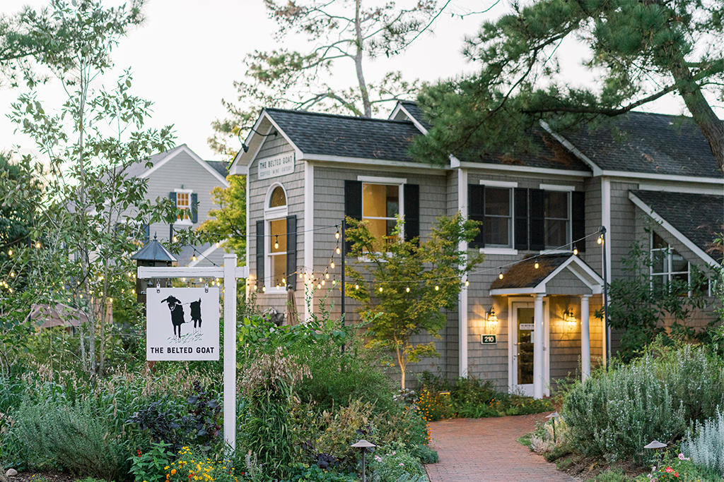 Charming two-story house with a sign that says "The Belted Goat" in front. Surrounded by lush greenery and trees, with string lights adorning the garden path leading to the entrance. Fearrington Village