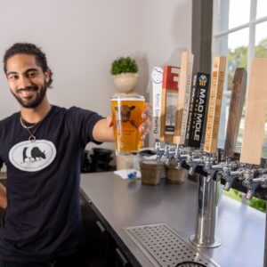 A man with a beard, wearing a black shirt with a bear logo, smiles while holding a glass of beer in front of a row of taps. The taps have various logos and labels. There's a window in the background with a view of greenery. Fearrington Village