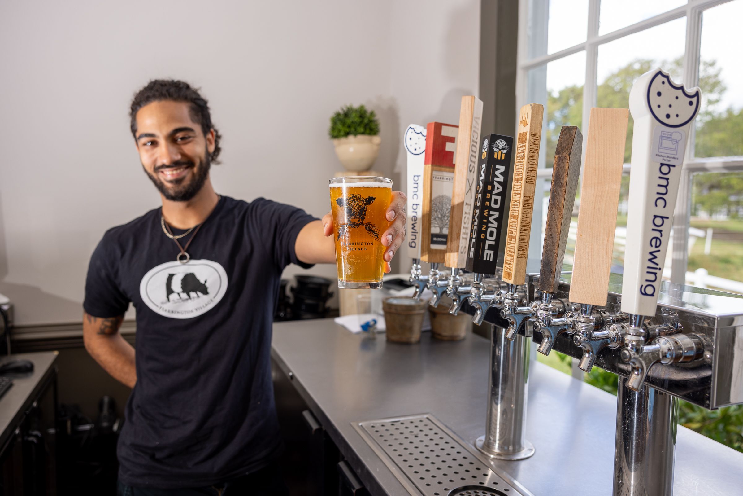 A man with a beard, wearing a black shirt with a bear logo, smiles while holding a glass of beer in front of a row of taps. The taps have various logos and labels. There's a window in the background with a view of greenery. Fearrington Village