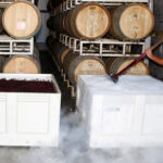 A person is standing next to large white bins filled with grapes, surrounded by fog, in a wine storage facility. Stacks of oak barrels line the background. The person is using a tool to work with the grapes. Fearrington Village