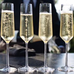 Four flutes of champagne are aligned on a table outdoors, with bubbles rising in each glass. A blurred background shows greenery and a person in dark formal attire. Fearrington Village