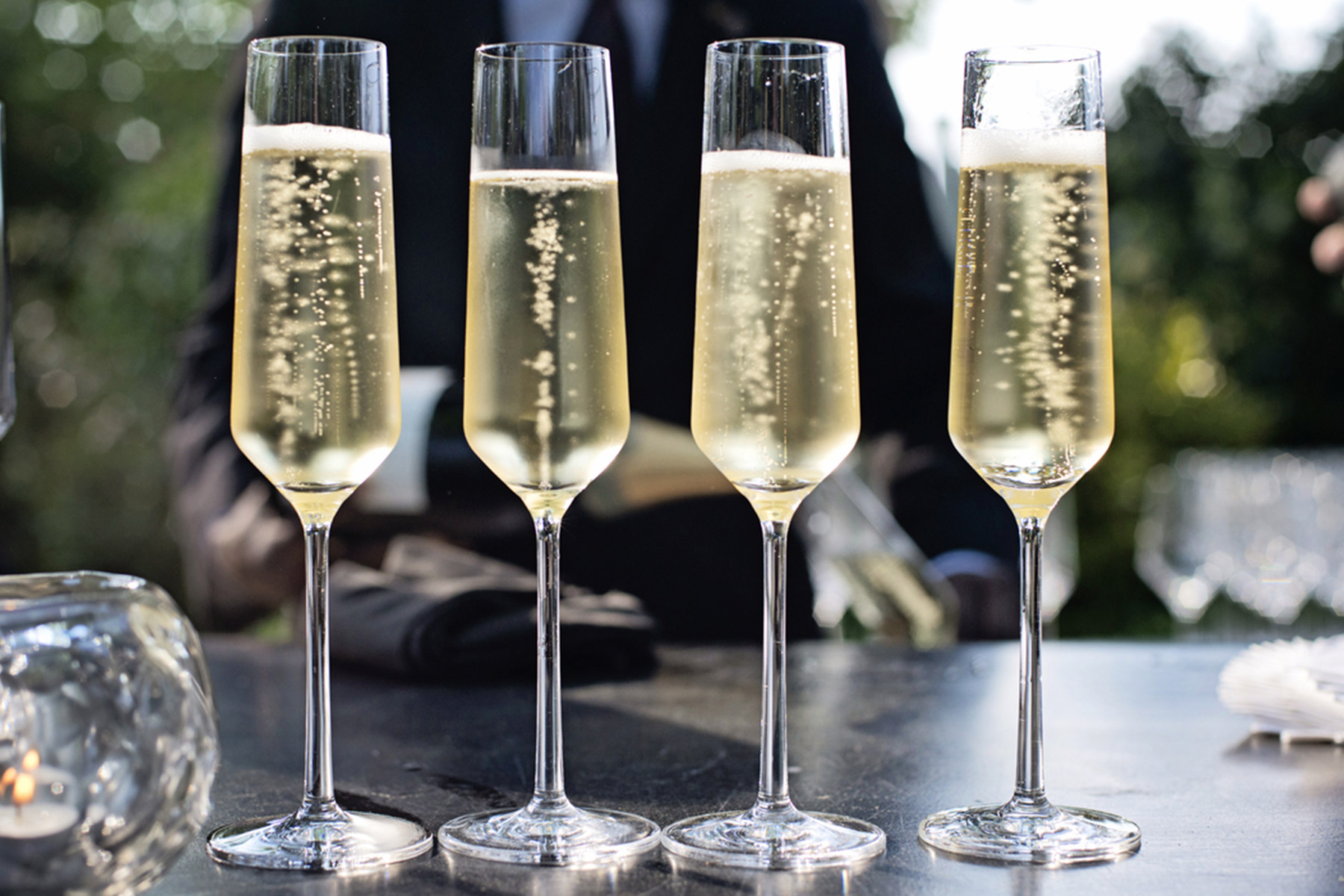 Four flutes of champagne are aligned on a table outdoors, with bubbles rising in each glass. A blurred background shows greenery and a person in dark formal attire. Fearrington Village