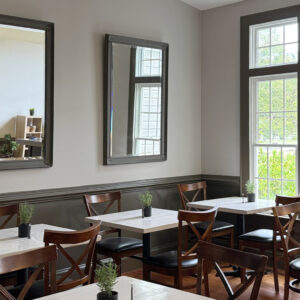 A cozy cafe interior with wooden chairs and white tables. Each table has a small potted plant. Large mirrors adorn the gray walls, and natural light streams in through tall windows, illuminating the space. Fearrington Village