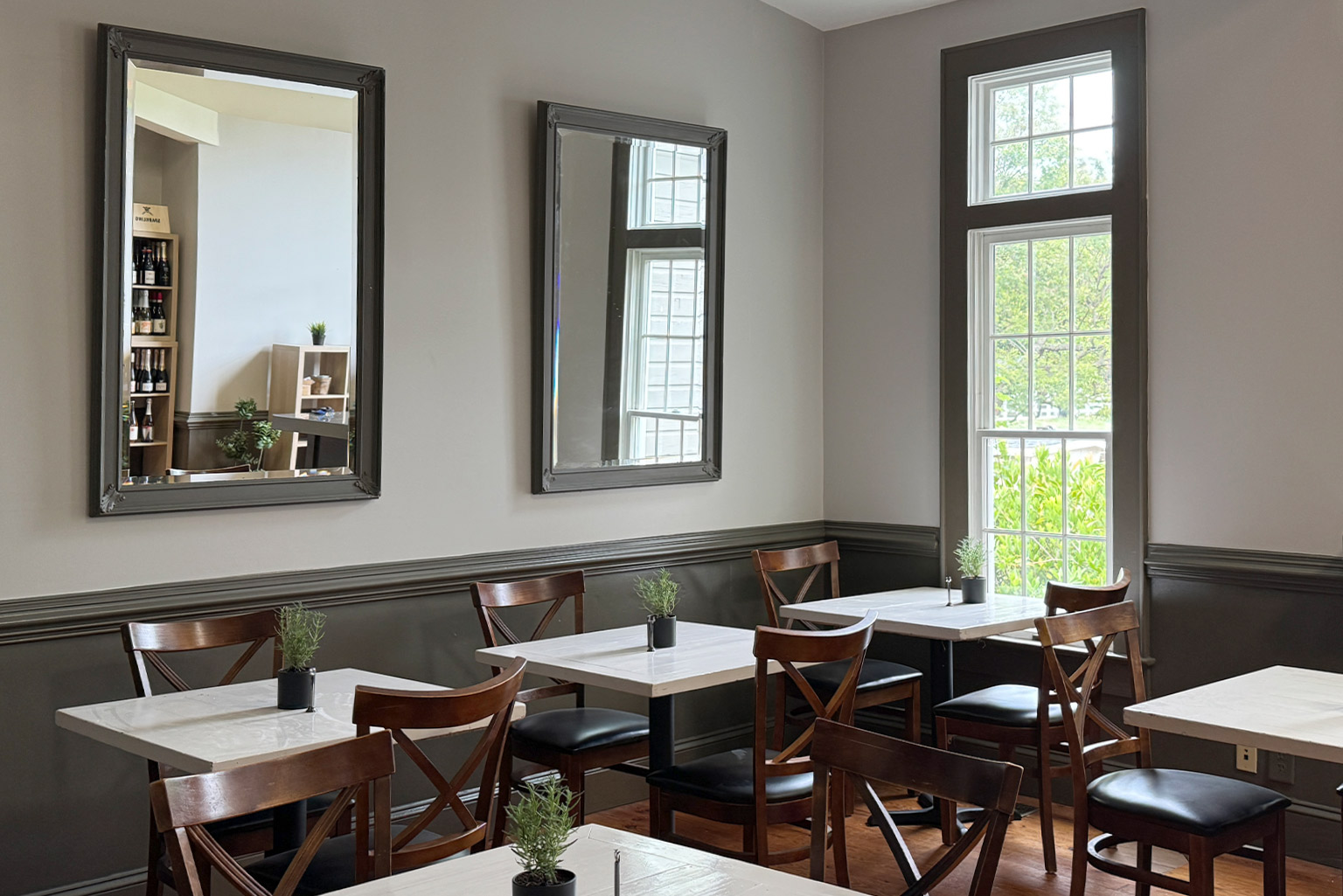 A cozy cafe interior with wooden chairs and white tables. Each table has a small potted plant. Large mirrors adorn the gray walls, and natural light streams in through tall windows, illuminating the space. Fearrington Village