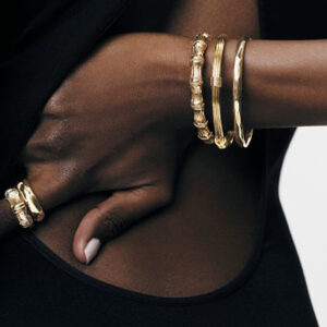 A close-up of a person wearing a black sleeveless outfit. Three gold bangles adorn their wrist, and two matching gold rings are on their fingers. The background is plain and light-colored. Fearrington Village