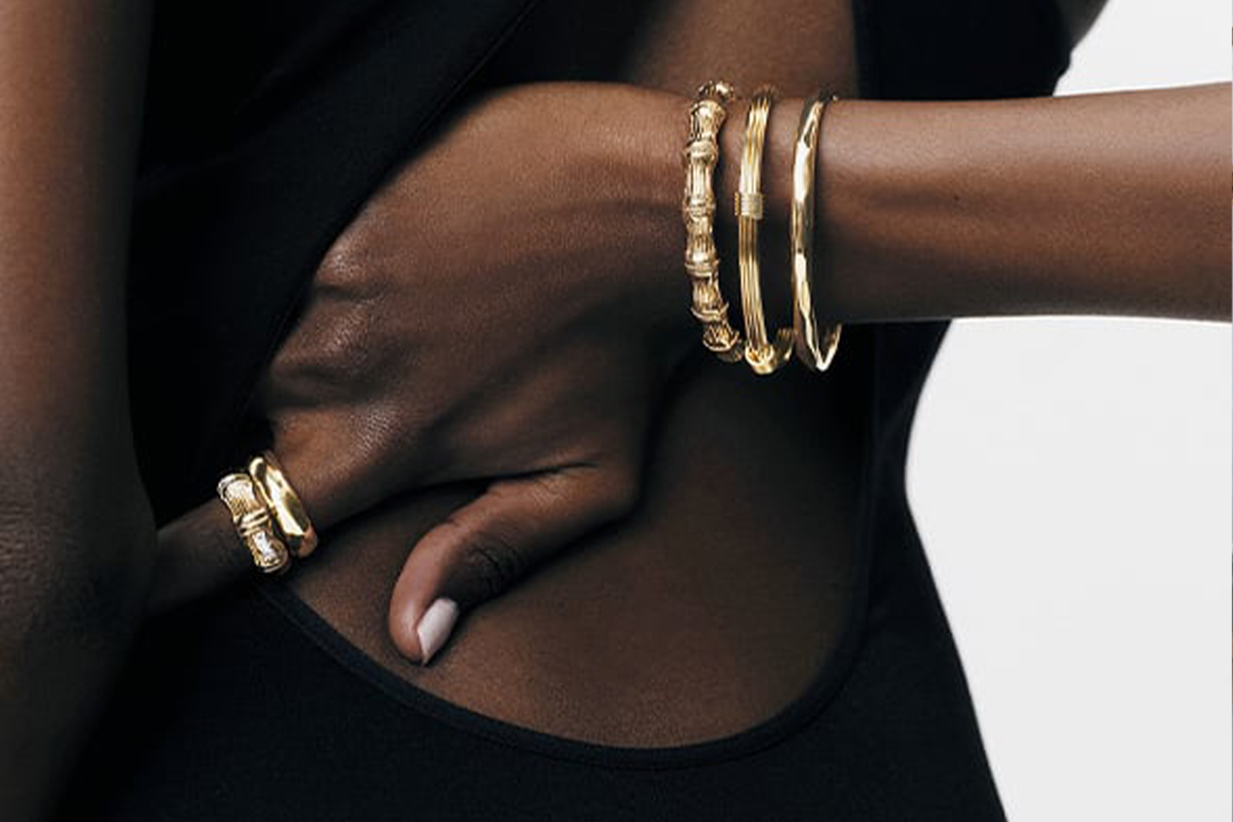 A close-up of a person wearing a black sleeveless outfit. Three gold bangles adorn their wrist, and two matching gold rings are on their fingers. The background is plain and light-colored. Fearrington Village