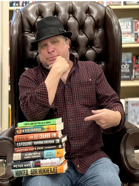 A person in a fedora and checkered shirt sits in a leather chair, pointing at a stack of books on their lap. The shelf behind them is filled with more books, suggesting a bookstore or library setting. Fearrington Village