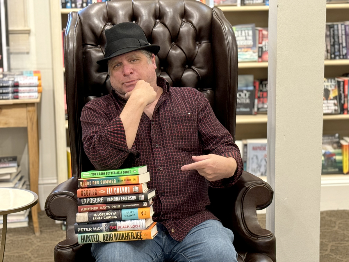 A man in a black hat and checkered shirt sits in a leather chair at a bookstore, pointing to a stack of books on a table. The background features bookshelves filled with various titles. Fearrington Village