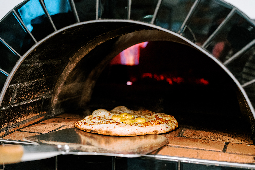 A pizza with melted cheese and toppings is being baked in a traditional wood-fired oven. A pizza peel is partially visible in the foreground, ready to remove the pizza. The oven's flames and brick interior are in the background. Fearrington Village