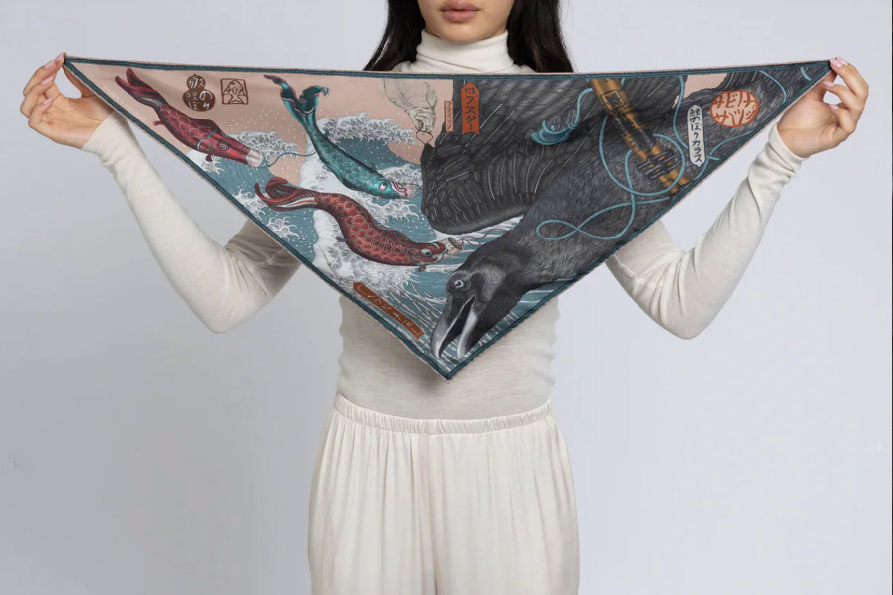 A person in a white turtleneck holds a triangular scarf featuring a design of a crow, waves, and red and teal-colored fish with Japanese-style artwork elements and symbols. Fearrington Village