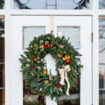 A festive holiday wreath adorned with red berries, dried orange slices, and a cream-colored ribbon hangs on a white door. The door features rectangular and circular glass panels, reflecting the soft glow of lights behind it. Fearrington Village