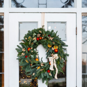 A festive holiday wreath adorned with red berries, dried orange slices, and a cream-colored ribbon hangs on a white door. The door features rectangular and circular glass panels, reflecting the soft glow of lights behind it. Fearrington Village