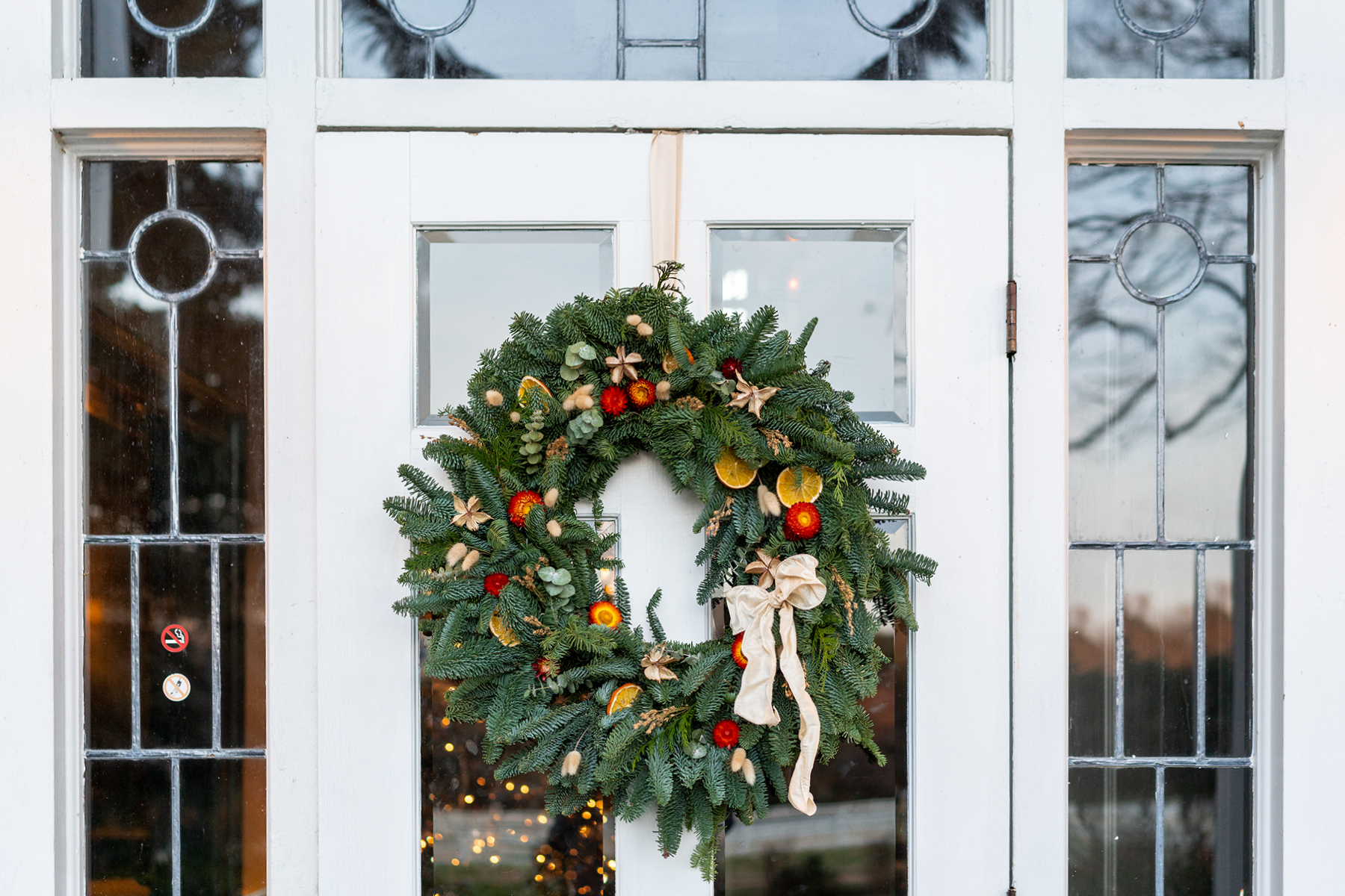 A festive holiday wreath adorned with red berries, dried orange slices, and a cream-colored ribbon hangs on a white door. The door features rectangular and circular glass panels, reflecting the soft glow of lights behind it. Fearrington Village