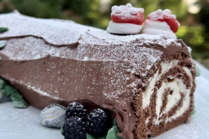 A Yule log cake with chocolate frosting, cream spirals, and powdered sugar. It is decorated with two small mushroom-shaped meringues on top and a few blackberries and green leaves at the base. Fearrington Village