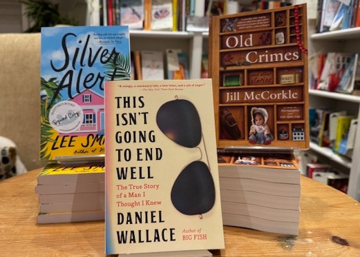 Three stacks of books displayed on a round wooden table in a bookstore. The visible titles are "Silver Alert" by Lee Smith, "This Isn't Going to End Well" by Daniel Wallace, and "Old Crimes" by Jill McCorkle. Shelves filled with books are in the background. Fearrington Village