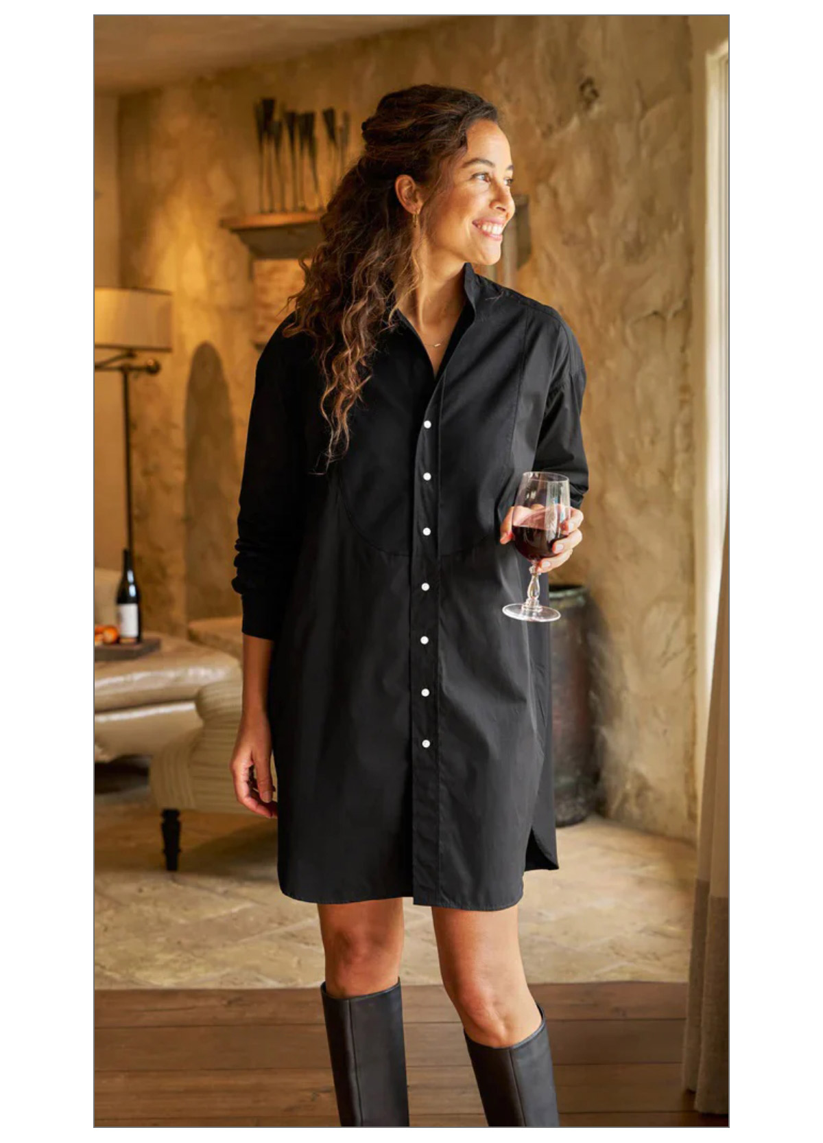 A woman smiles while standing indoors, wearing a black button-up shirt dress and holding a glass of red wine. The room has rustic decor, including a fireplace and a bottle of wine on a table in the background. Fearrington Village