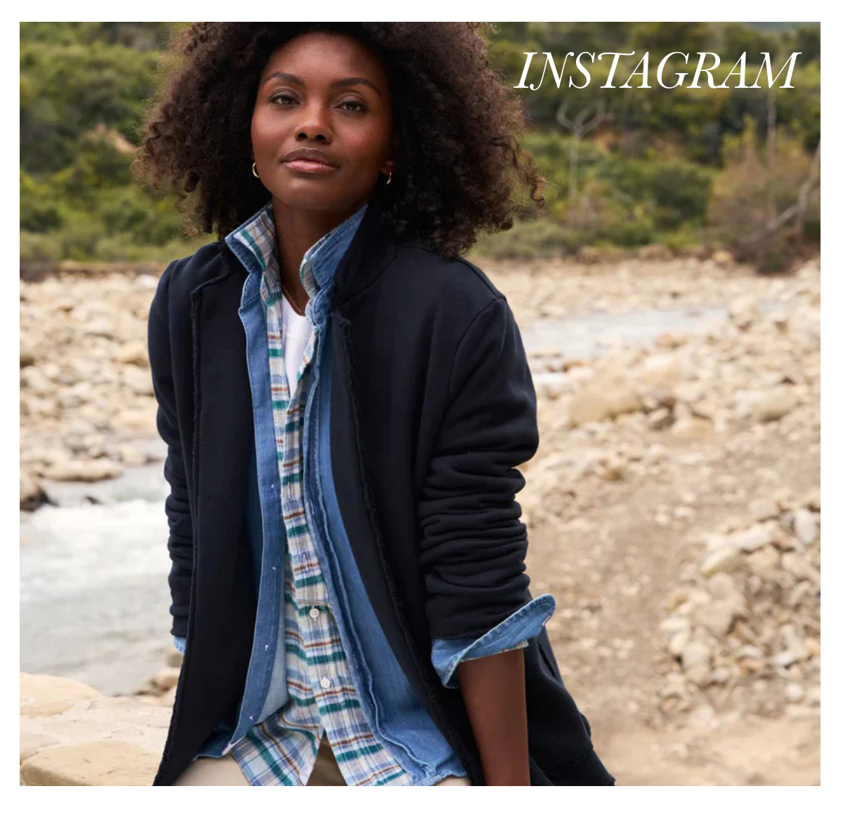 A person with curly hair wearing a layered outfit with a plaid shirt and dark jacket stands by a rocky riverside. The background shows trees and a flowing stream. The word "INSTAGRAM" is written in the top right corner. Fearrington Village