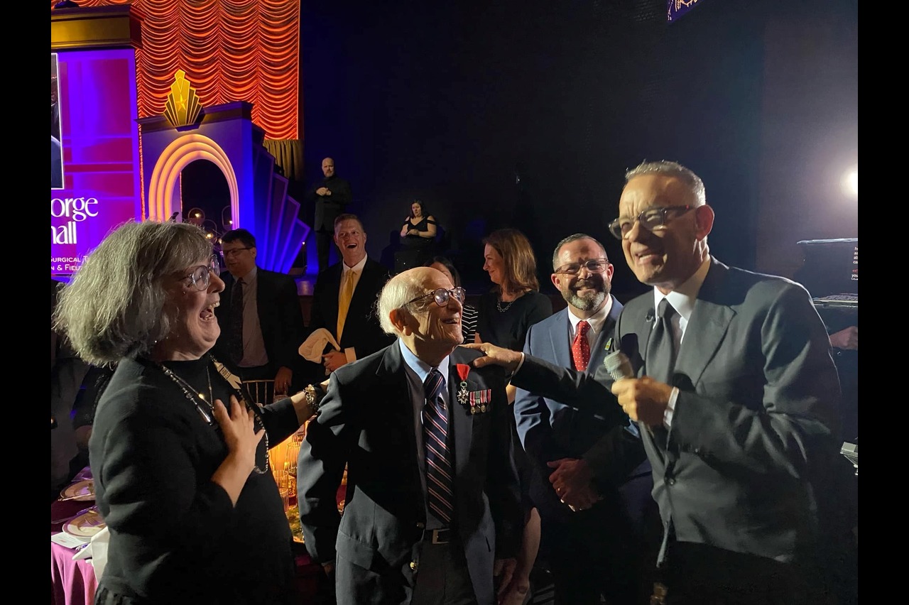 A lively event with a group of people engaged in conversation. A man in a suit holds a microphone, smiling, while others around him, including an elderly man with medals, laugh and engage. The setting is warmly lit with vibrant stage decorations. Fearrington Village