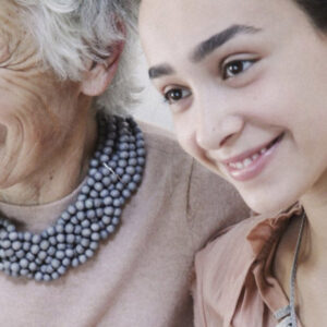 An elderly woman with a beaded necklace and a younger woman are smiling together. The background is softly blurred, focusing on their joyful expressions and close bond. Fearrington Village