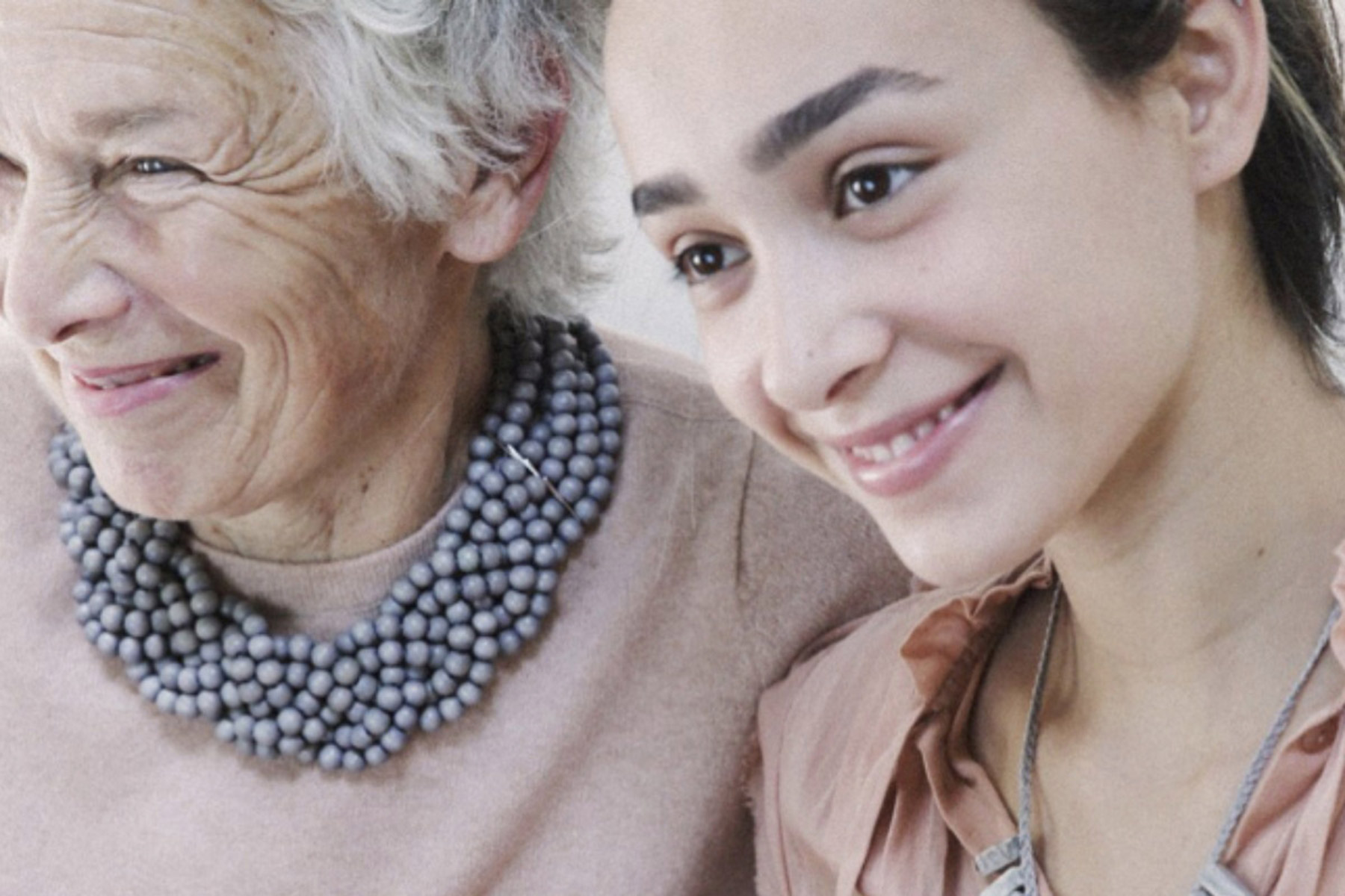 An elderly woman with a beaded necklace and a younger woman are smiling together. The background is softly blurred, focusing on their joyful expressions and close bond. Fearrington Village