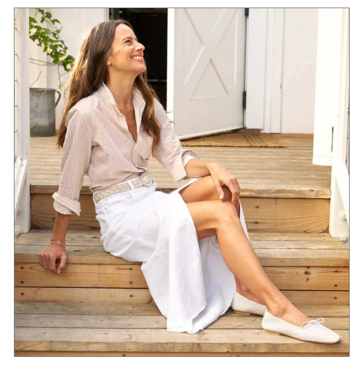 A woman with long hair and a striped shirt smiles while sitting on wooden steps. She wears a white skirt and shoes, with a relaxed and content expression, outdoors near an open door and potted plant. Fearrington Village