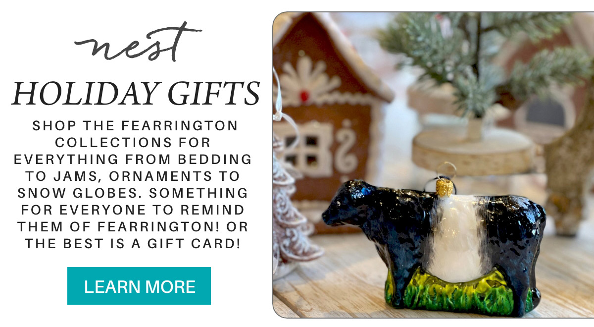 Holiday gift display featuring a cow ornament in front of gingerbread house decorations and a small tree. Includes text promoting Fearrington collections with items like bedding, jams, and ornaments. A "Learn More" button is included. Fearrington Village