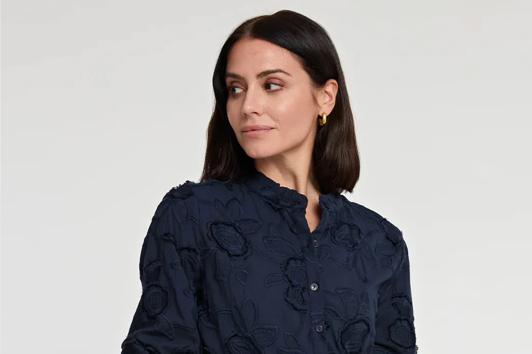 A woman with shoulder-length dark hair wears a navy blouse with floral embroidery. She is looking to her left against a plain light background. Fearrington Village