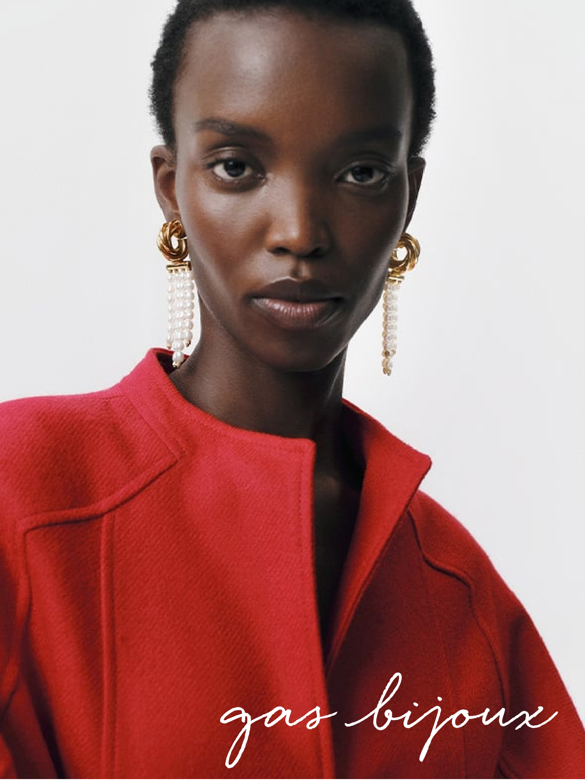 A person with short hair wears a vibrant red jacket and large, ornate gold and pearl earrings. The text "gas bijoux" is written in elegant white cursive at the bottom of the image. Fearrington Village