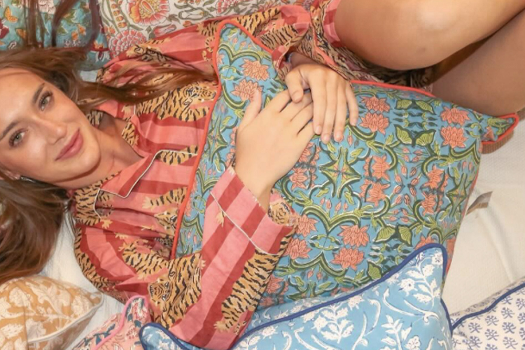 A person in patterned pajamas lies on a bed, surrounded by colorful, floral pillows. They are holding a pillow with a blue and pink floral design, and their hair is spread out across the bedding. Fearrington Village