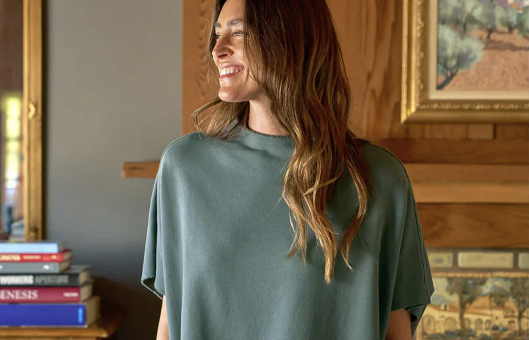 A woman with long brown hair smiling and wearing a loose green top stands in a warmly lit room. She is near a fireplace with a wooden mantle, a stack of books, and a framed painting hanging on the wall. Fearrington Village